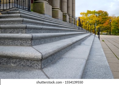 The Corner Of Granite Steps To A Large Historic Government Building.The Court House Has Large Columns.  There's A Black Metal Handrail Leading Up To The Building. Autumn Trees Are In The Background.  