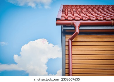 Corner of the house with metal tile roof and rain gutter. Red gutter on the roof top of house. Rooftop water collection system. Metallic Guttering System, Guttering and Drainage Pipe Exterior