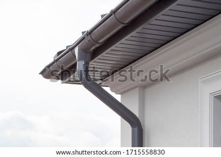 Corner of the house with new gray metal tile roof and rain gutter. Metallic Guttering System, Guttering and Drainage Pipe Exterior