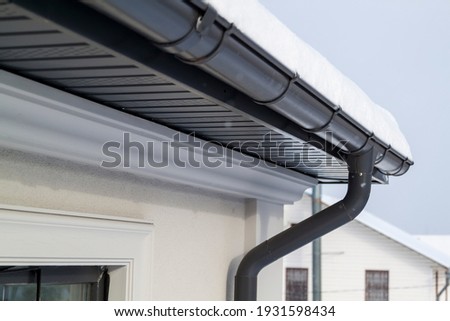 Corner of the house with new gray metal tile roof and rain gutter at winter. Metallic Guttering System, Guttering and Drainage Pipe Exterior