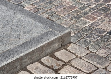 Corner Of A Pedestrian Sidewalk Made Of Granite Curb And Stone Rectangular Brick Tiles Nearby A Road Paved With Rough Cut Paving Slabs Close-up, Nobody.