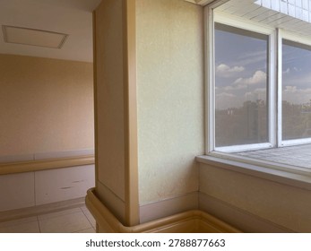 Corner perspective of an empty bright institutional corridor featuring beige walls, safety handrails, and large glass windows revealing a sunny sky with fluffy white clouds and distant trees.