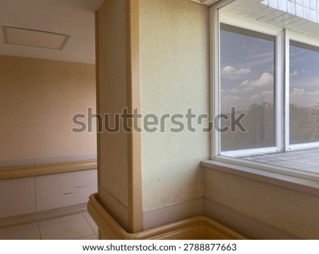 Corner perspective of an empty bright institutional corridor featuring beige walls, safety handrails, and large glass windows revealing a sunny sky with fluffy white clouds and distant trees.