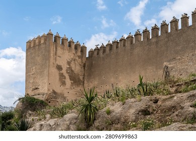 The corner rampart tower and a long section of the ancient defensive walls of Fes el-Bali near Bab Mahrouk show crenellated battlements and pise construction against a blue sky with clouds.
