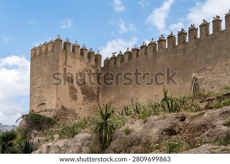 The corner rampart tower and a long section of the ancient defensive walls of Fes el-Bali near Bab Mahrouk show crenellated battlements and pise construction against a blue sky with clouds.