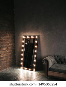 Corner room with a wooden and  gray cement wall, an aged floor, black glass doors and electric dressing makeup mirror, lights bulbs tern on, sofa. Loft style