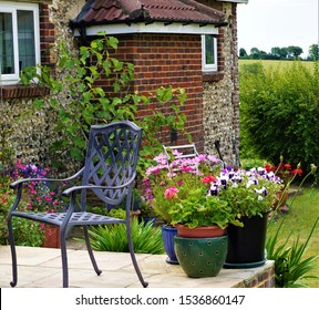 Corner Of A Sussex Flint Farmhouse And Garden Featuring Metal Patio Chair And Pots Of Flowers In Foreground.