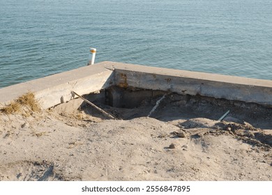 Corner view of eroded storm damaged seawall. Cracks in the seawall vertical panels and cap. Calm Blue bay water in the back on a sunny day. White and red navigation buoy.  

