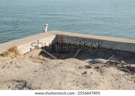 Corner view of eroded storm damaged seawall. Cracks in the seawall vertical panels and cap. Calm Blue bay water in the back on a sunny day. White and red navigation buoy.  
