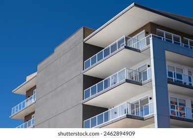 Corner view of a new apartment complex. The wall is grey modern metal paneling. Each unit has a glass balcony with a glass patio door. The condo units are on the corner of the modern style building. 