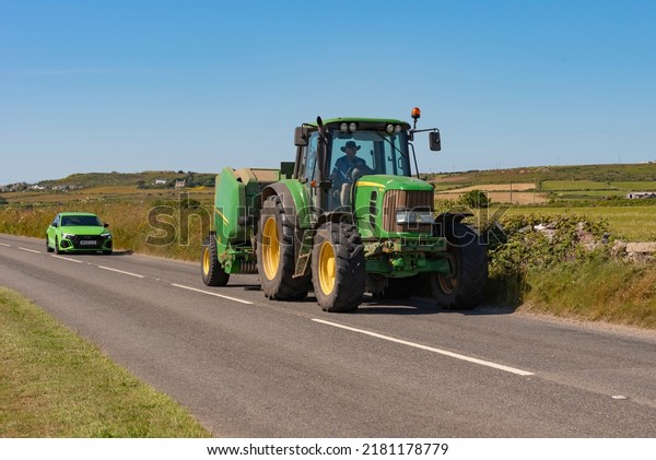 Cornwall, England, UK. 2022. Green car following a\
green tractor and trailer travelling along a country lane close to\
St Just, Cornwall, UK