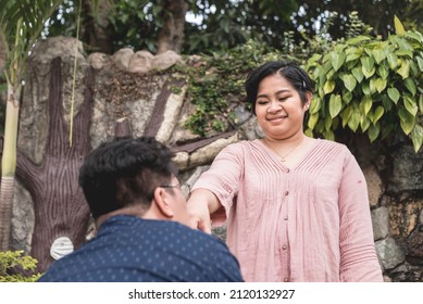 A Corny Man Kisses The Ring Finger Of His Young Filipino Wife. Proclaiming Love In A Mushy Way. Outdoor Garden Scene.