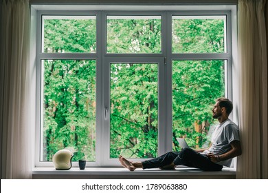 Corona virus - staying and work at home (self-isolation, self-quarantine). A self-isolated handsome man sits alone in front of a panoramic window with a laptop. Online shopping, home work, freelance.
