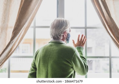 Coronavirus. Back View Of A Senior White Haired Man With Green Sweater In Solitude At Home Behind The Window, Wearing A Protective Face Mask Due To Coronavirus Infection - Elderly People In Quarantine