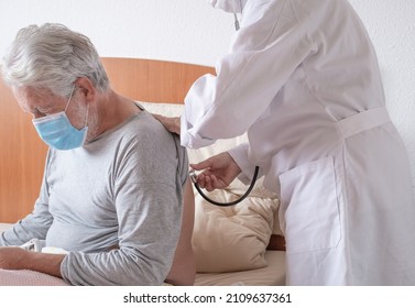 Coronavirus covid 19. Doctor listening to an elderly man's breathing in bed during a home visit. Elderly white-haired patient wearing mask infected with sars-cov 2 omicron variant in home quarantine - Shutterstock ID 2109637361