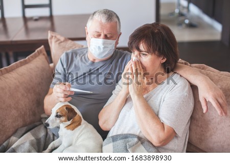 Coronavirus CoVid-19 Couple old aged senior people at home with seasonal winter cold illness disease sit down on the sofa. Elderly couple in medical masks during the pandemic