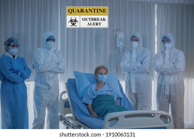  Coronavirus Covid-19 Infected Senior Caucasian Patient On Hospital Bed , Doctor And Medical Team In PPE Coverall Suit Standing By Arm Cross  In Quarantine Room And Outbreak Alert Sign.