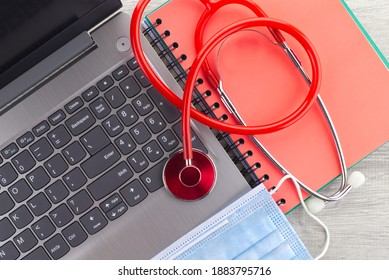 Coronavirus Or Covid-19 Medical Concept With A Colorful Red Stethoscope Coiled Over A Surgical Face Mask Alongside A Notebook And Pen On A Wooden Desk With Computer Keyboard Viewed Top Down