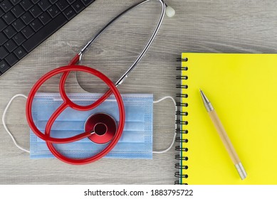 Coronavirus Or Covid-19 Medical Concept With A Colorful Red Stethoscope Coiled Over A Surgical Face Mask Alongside A Notebook And Pen On A Wooden Desk With Computer Keyboard Viewed Top Down