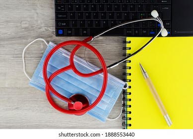 Coronavirus Or Covid-19 Medical Concept With A Colorful Red Stethoscope Coiled Over A Surgical Face Mask Alongside A Notebook And Pen On A Wooden Desk With Computer Keyboard Viewed Top Down