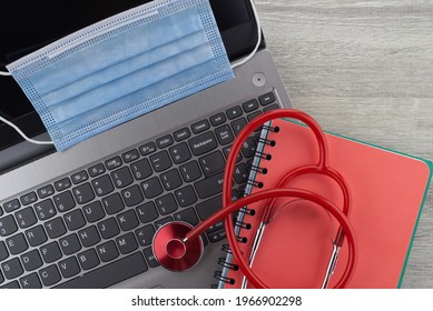 Coronavirus Or Covid-19 Medical Concept With A Colorful Red Stethoscope Coiled Over A Surgical Face Mask Alongside A Notebook And Pen On A Wooden Desk With Computer Keyboard Viewed Top Down