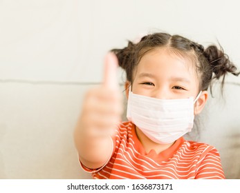 Coronavirus Covid-19.Online Education.Little Asian Kid Girl Wearing Face Mask Show Thumbs Up For Thank You Doctor, Happy At Home. Covid-19 Coronavirus.Stay Home.Social Distancing.New Normal Behavior.