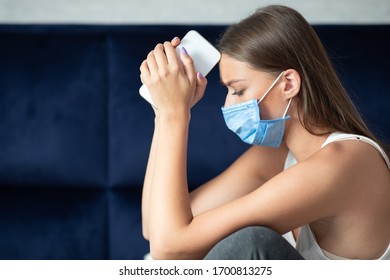 Coronavirus And Depression. Unhappy Woman In Medical Mask Holding Cellphone Sitting On Sofa At Home During Corona Virus Quarantine.