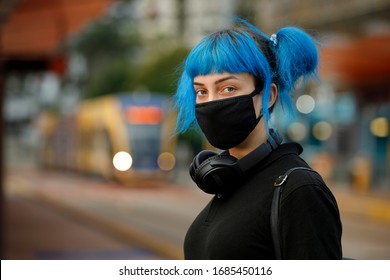 Coronavirus Fashionable Medical Face Mask Worn By Young Female Student With Blue Anime Style Hair, Standing At Tram Stop On A Urban City Street On Dusk, Stop Covid 19 Pandemic Or Air Pollution Concept