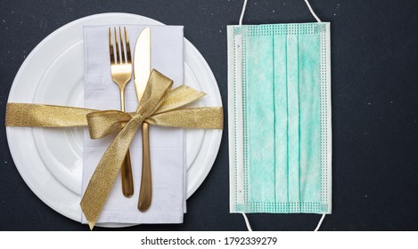 Coronavirus And Holiday Table Concept. Face Protective Mask And Festive Golden Table Setting Against Black Color Background. COVID Spread Prevention Measure