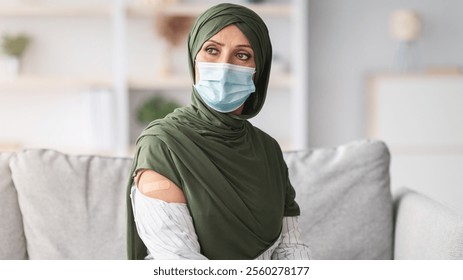 Coronavirus Immunization Campaign. Vaccinated Muslim Female Showing Arm With Medical Plaster Bandage After Antiviral Vaccination Sitting On Couch At Home, Wearing Hijab And Protective Face Mask - Powered by Shutterstock