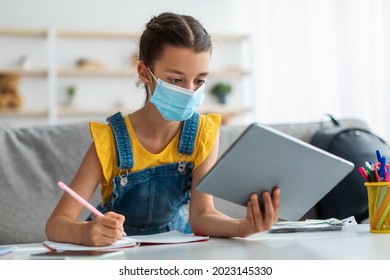 Coronavirus Impact On Education. Girl Teenager Wearing Medical Mask Studying Online Using Digital Tablet, Writing Notes In Copybook, Free Space. Female Student Learning Remotely During Covid Outbreak