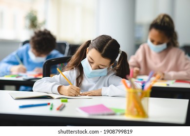 Coronavirus Impact On Education. Little Girl Sitting At Table In Classroom At School Or Kindergarten, Wearing Protective Medical Face Mask, Writing Or Drawing In Textbook. New Rules And Protection