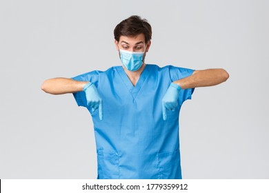 Coronavirus Outbreak, Healthcare Workers Fighting Disease, Hospitals Concept. Curious And Excited Doctor Blue Scrubs, Gloves And Medical Mask, Pointing Fingers Down, Look Interested At Advertisement