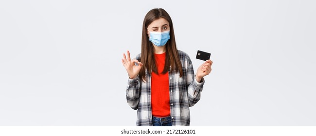 Coronavirus Outbreak, Working From Home, Online Shopping And Contactless Payment Concept. Pleased Young Female Bank Customer In Medical Mask, Show Okay Sign And Credit Card, Wink