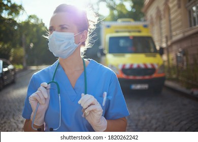 Coronavirus Pandemic. Modern Paramedic Woman In Uniform With Stethoscope And Medical Mask Looking Into The Distance Outside Near Ambulance.