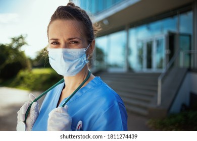 Coronavirus Pandemic. Portrait Of Smiling Modern Physician Woman In Scrubs With Stethoscope And Medical Mask Outdoors Near Clinic.