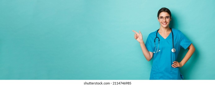 Coronavirus, pandemic and social distancing concept. Image of smiling confident doctor in glasses and scrubs, pointing fingers left at logo on copy space, standing over blue background