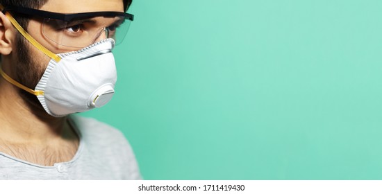 Coronavirus Prevention. Close-up Studio Portrait Of Young Guy With Medical Respiratory Mask Against Flu, Wearing Safety Goggles, On Background Of Aqua Menthe Color.