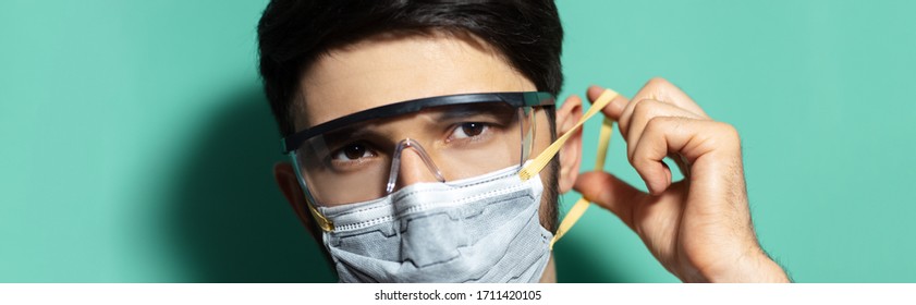 Coronavirus Prevention. Panoramic Portrait Of Young Guy Put On Medical Respiratory Mask, Wearing Safety Goggles On Background Of Aqua Menthe Color.