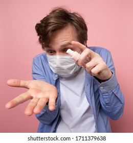 Coronavirus Protection And Disinfection Or OCD Concept. Caucasian Man In Medical Mask Spraying Sanitizer Spray On His Hand Over Pink Background