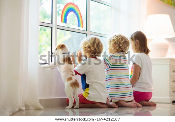 Coronavirus quarantine. Stay home. Kids sitting at window. Children drawing rainbow sign of hope. Boy and girl during corona virus lockdown. Child and pet. Family isolation indoors. Disease prevention