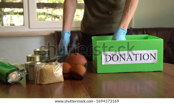 Coronavirus\
Relief Funds and Donations. Volunteer in the Protective Medical\
Mask and hand Gloves Putting Food In Donation Box. Charity\
donations. Making Donations To Food\
Bank