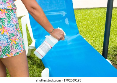 Coronavirus Safety Measures: Woman Using A Bottle Of Disinfectant To Clean A Sunbed In A Public Pool. Summer With Coronavirus And New Normal.