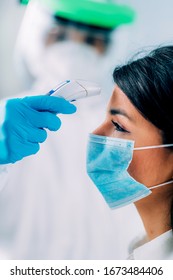 Coronavirus Screening. Medical Worker In Protective Suit Measuring Body Temperature With Contactless Body Thermometer, Young Woman Wearing Protective Mask