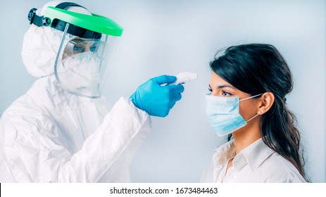 Coronavirus Screening. Medical Worker In Protective Suit Measuring Body Temperature With Contactless Body Thermometer, Young Woman Wearing Protective Mask