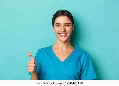 Coronavirus, social distancing and health concept. Close-up of cheerful female doctor, winking and smiling, showing thumbs-up in approval, standing upbeat over blue background - Shutterstock ID 1928494172