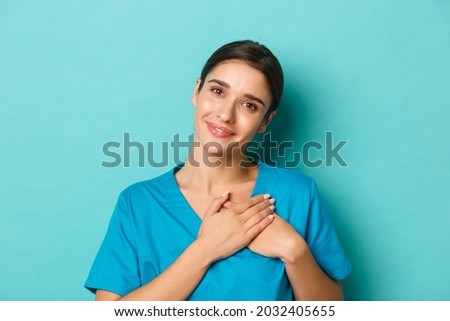 Coronavirus, social distancing and health concept. Close-up of smiling attractive female doctor, feeling grateful, holding hands on heart and looking with gratitude, standing over blue background