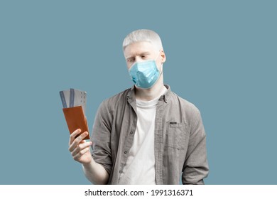 Coronavirus travel restrictions. Sad albino guy in face mask looking at passport and plane tickets, disappointed because of cancelled flight or closed borders, turquoise background - Powered by Shutterstock