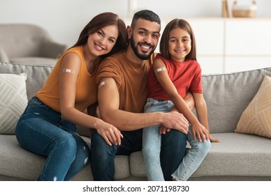 Coronavirus Vaccination Advert. Happy Vaccinated Family Of Three People Showing Arm With Sticking Plaster After Covid-19 Vaccine Vax Injection Posing Sitting On Couch In Living Room, Smiling To Camera