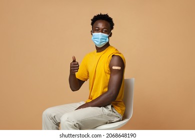 Coronavirus Vaccination Advertisement. Happy Vaccinated Black Guy With Plaster On Arm Showing Thumb Up After Getting Covid-19 Vaccine Injection, African Man In Medical Mask Posing On Beige Background